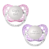 A set of two orthodontic pacifiers with silicone nipples, one in pink and one in purple, each engraved with the text 'Gigi's Girl' and  'Gig's Little Love'.