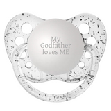 A clear silicone pacifier with 'MY GODFATHER LOVES ME' engraved on the front, along with a small round protective cap.