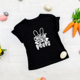 A black short-sleeved baby shirt with 'Chillin with my Peeps' text and bunny ears graphic, presented on a wooden surface with Easter-themed decorations around it.