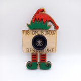 A wooden Christmas ornament designed as an elf with a red hat and a surveillance camera, with the text 'This Home is Under Elf Surveillance' engraved on it. The ornament is adorned with red and green details and a black camera lens.