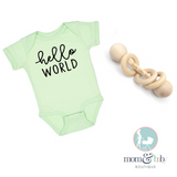 A green colored baby onesie with the text 'Hello World' printed on the front, alongside a wooden baby rattle.