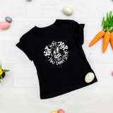 A black short-sleeved baby t-shirt with 'Hip to the Hop You Don't Stop' text and a graphic of a bunny, displayed on a wooden surface with Easter-themed decorations around it.